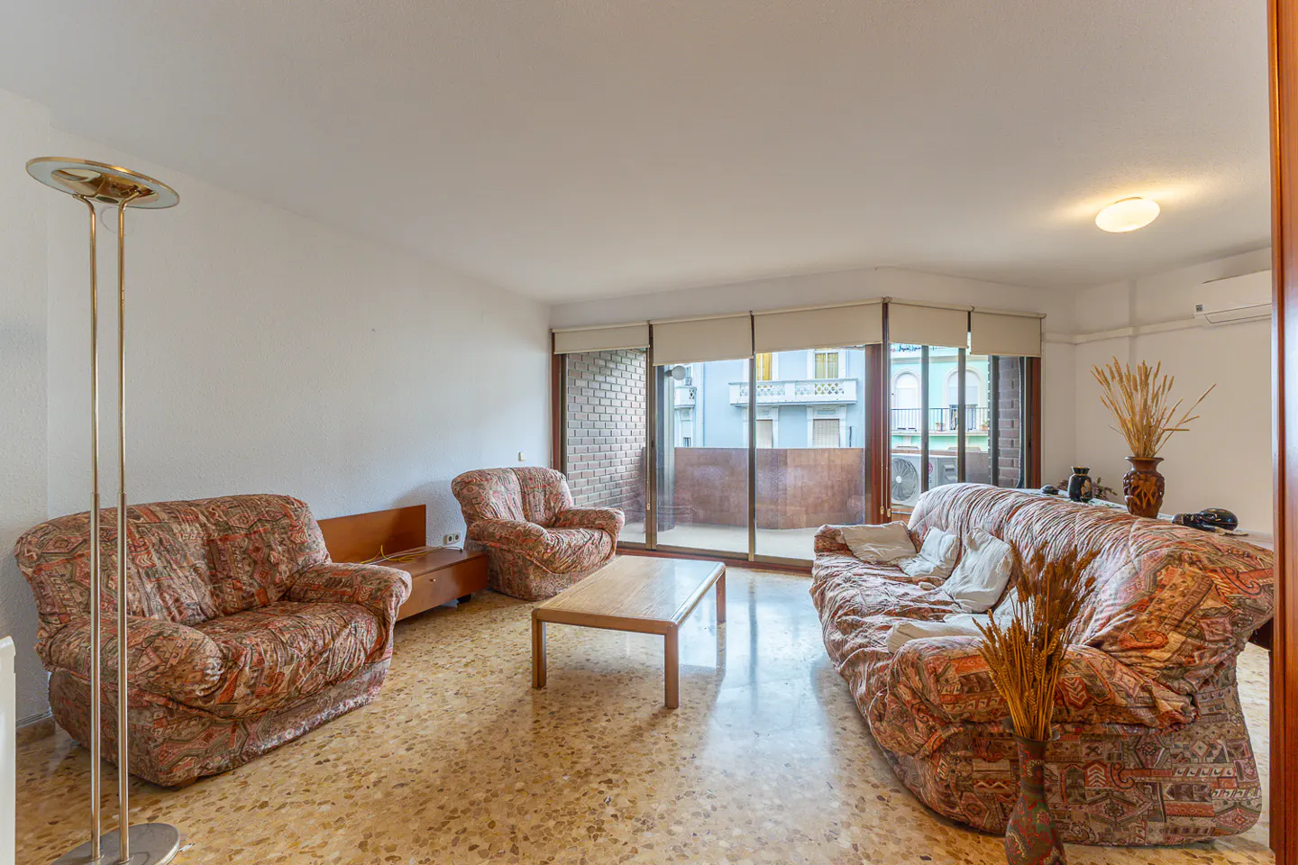 Living room with patterned sofas, a wooden coffee table, terrazzo floors, and a balcony view of city buildings.