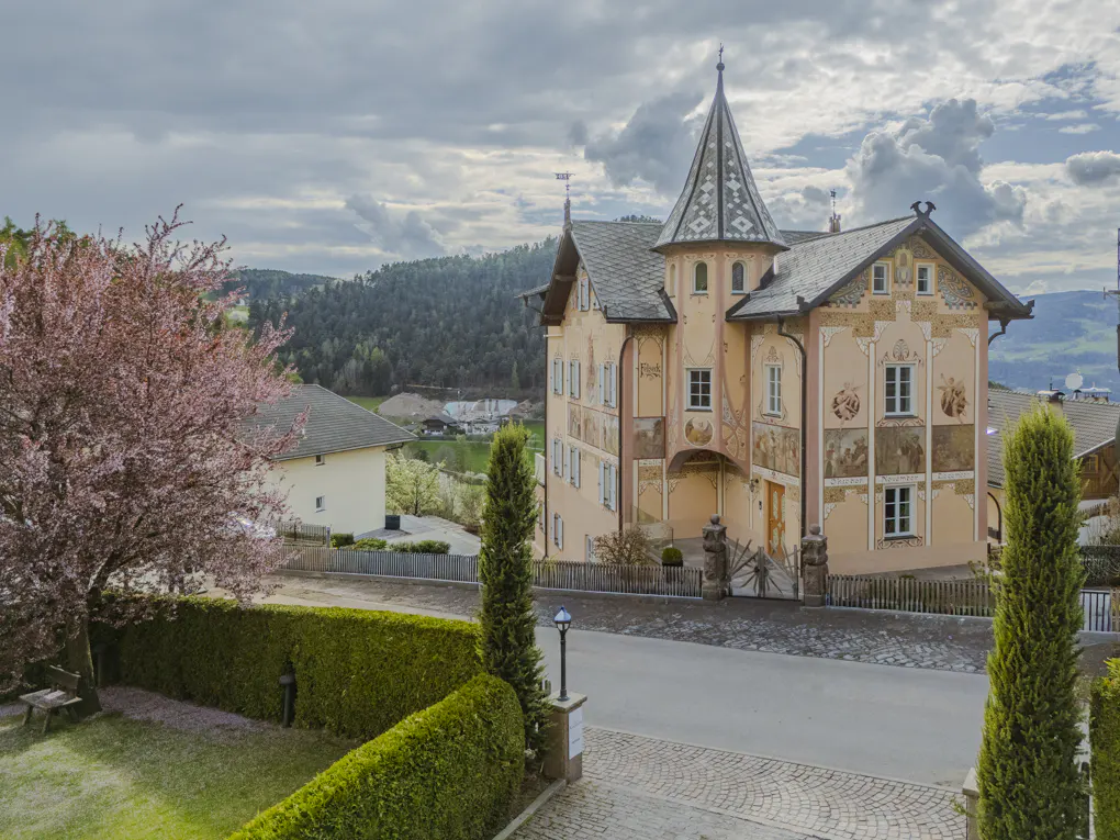 Exterior view of a peach-colored, ornate building with a turret, surrounded by greenery and a blooming pink tree.