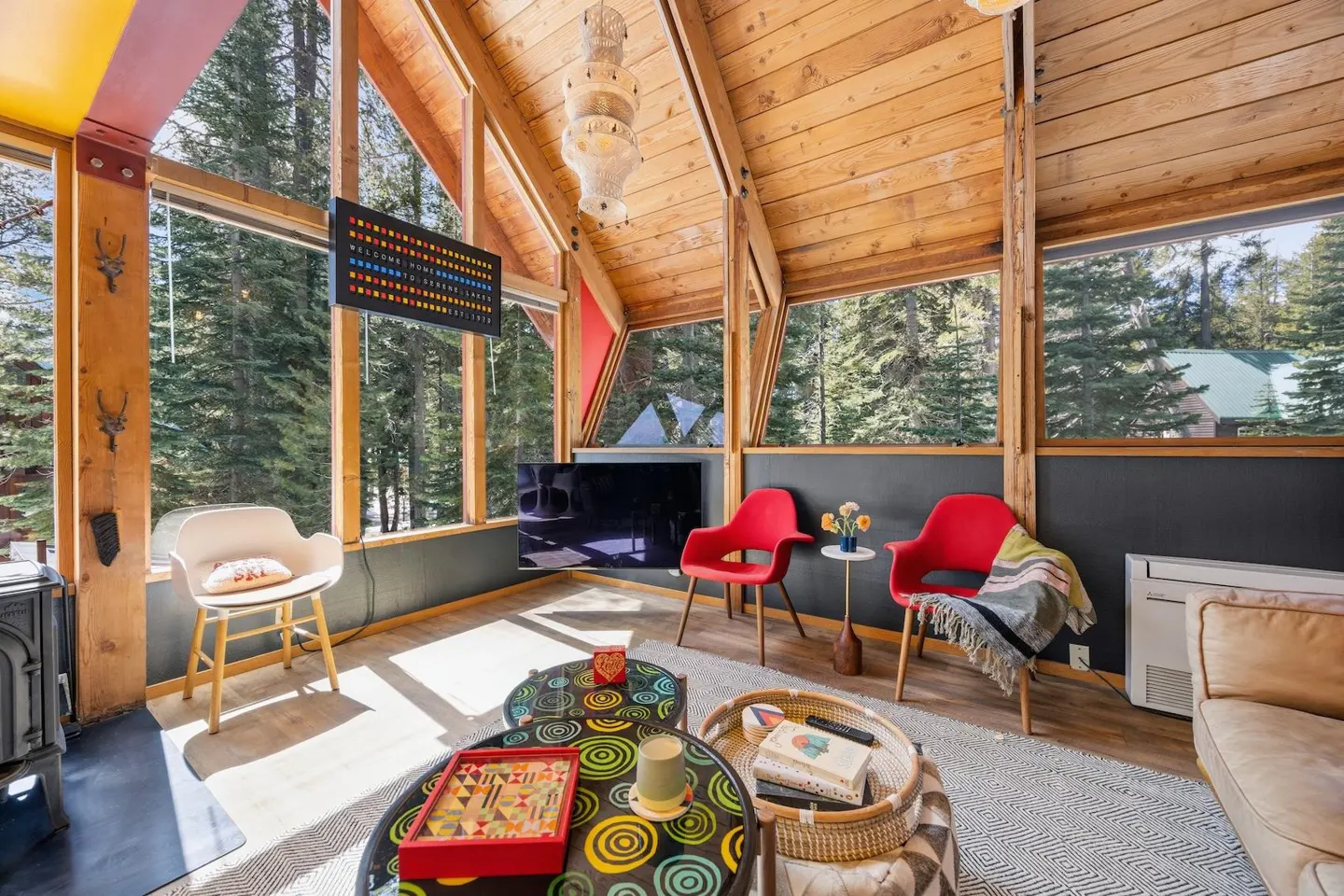 A-frame living room with wood ceiling, large windows with forest view, red chairs, and patterned coffee tables.