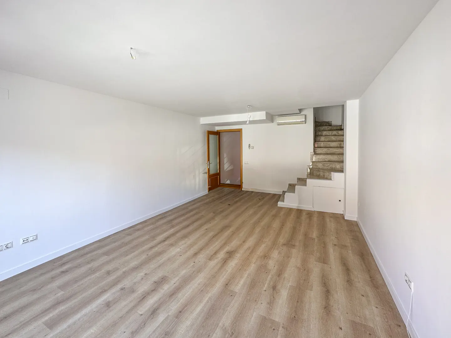 Empty, bright living room with white walls, light wood-look flooring, a wooden door, and stairs leading to an upper level.