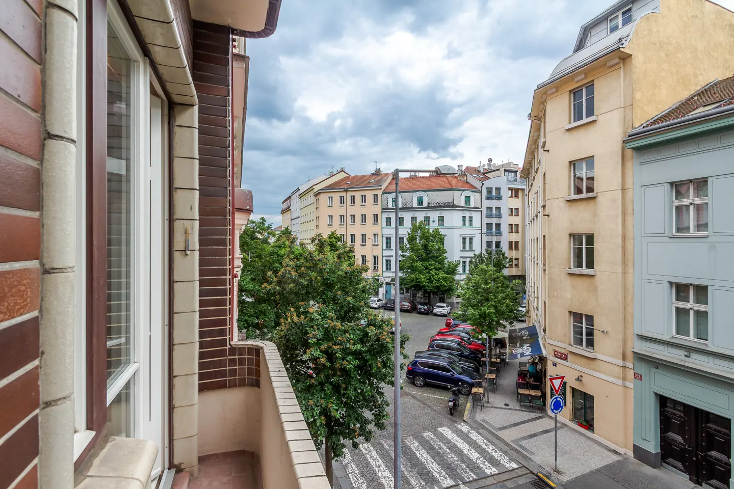 View from a balcony overlooking a European street with parked cars, trees, and buildings under a cloudy sky.