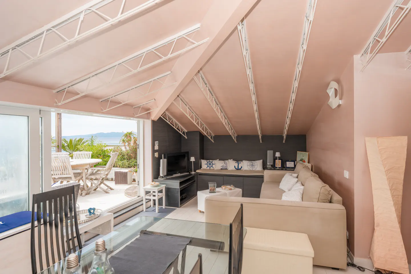 Attic living room with pink walls, white ceiling beams, and a beige sectional sofa. Sliding glass doors open to a patio with ocean views.