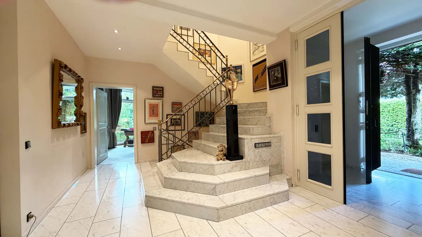 Elegant foyer with marble floors and stairs. A gold-framed mirror hangs on the left wall. Artwork adorns the stairwell.