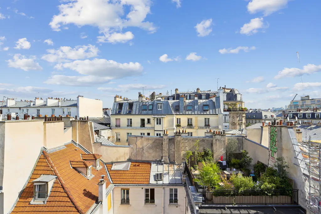Rooftop view of Paris buildings with orange tile roofs, chimneys, and a rooftop garden under a blue sky with white clouds.