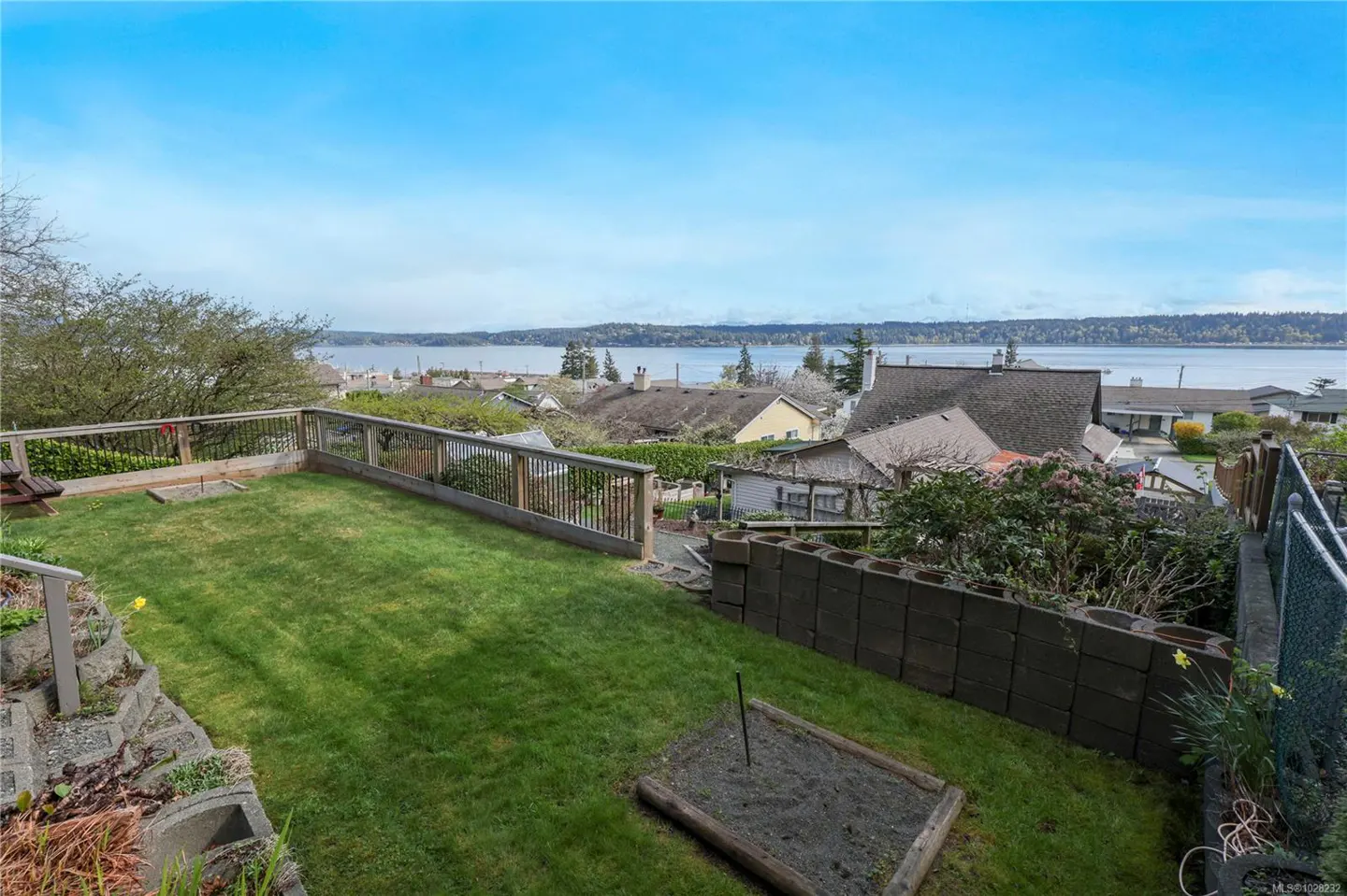 A backyard with green grass, a wooden fence, and a view of the ocean and houses in the distance.
