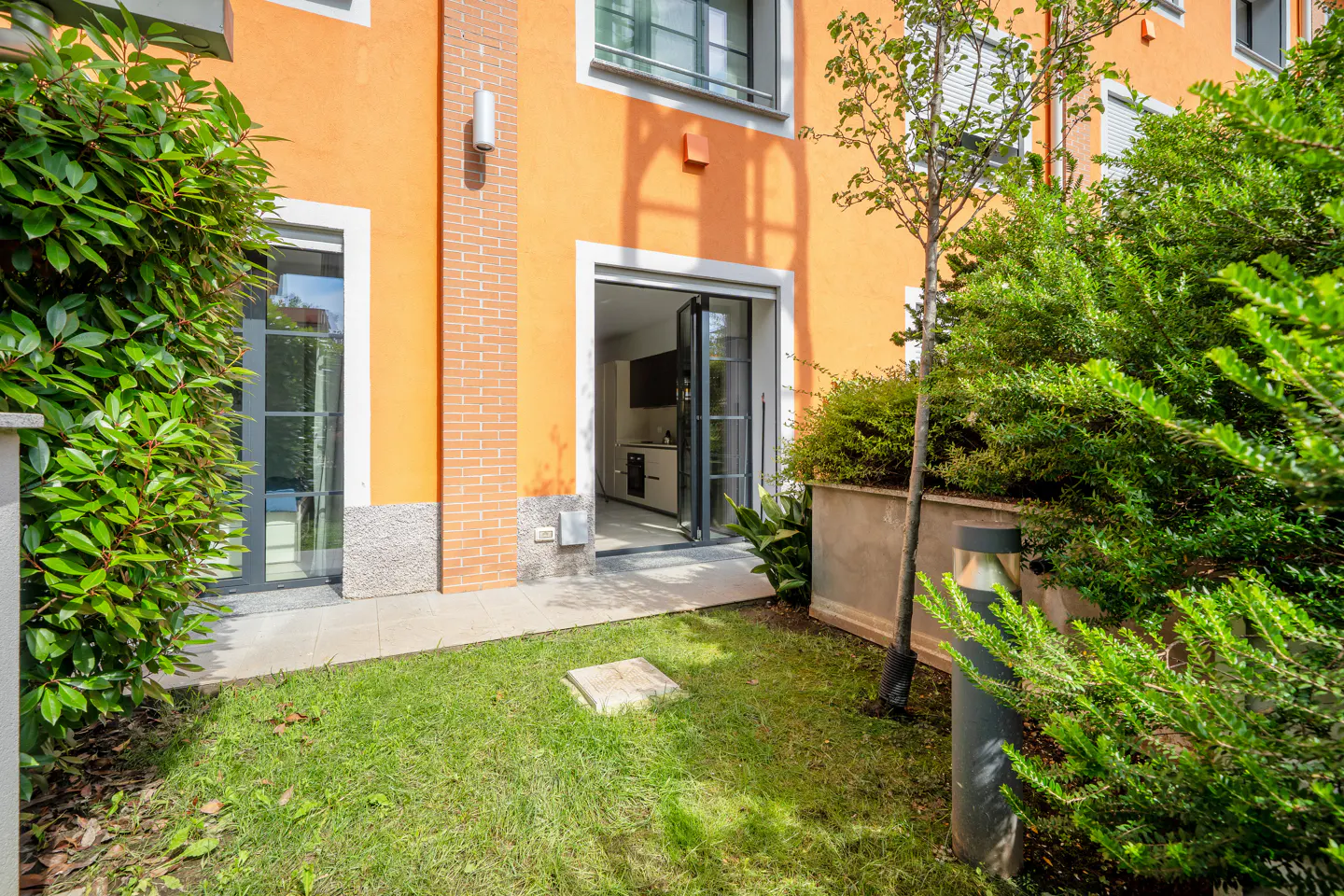 Small grassy yard with a modern orange building, glass doors opening to a kitchen, and lush green bushes.