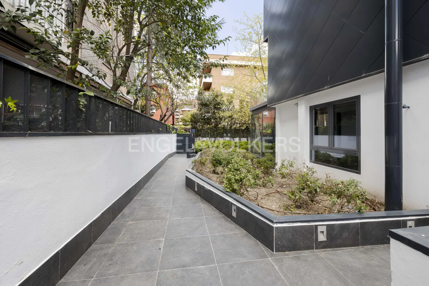 A paved walkway leads past a modern home with white walls, black metal accents, and a small garden bed with shrubs.