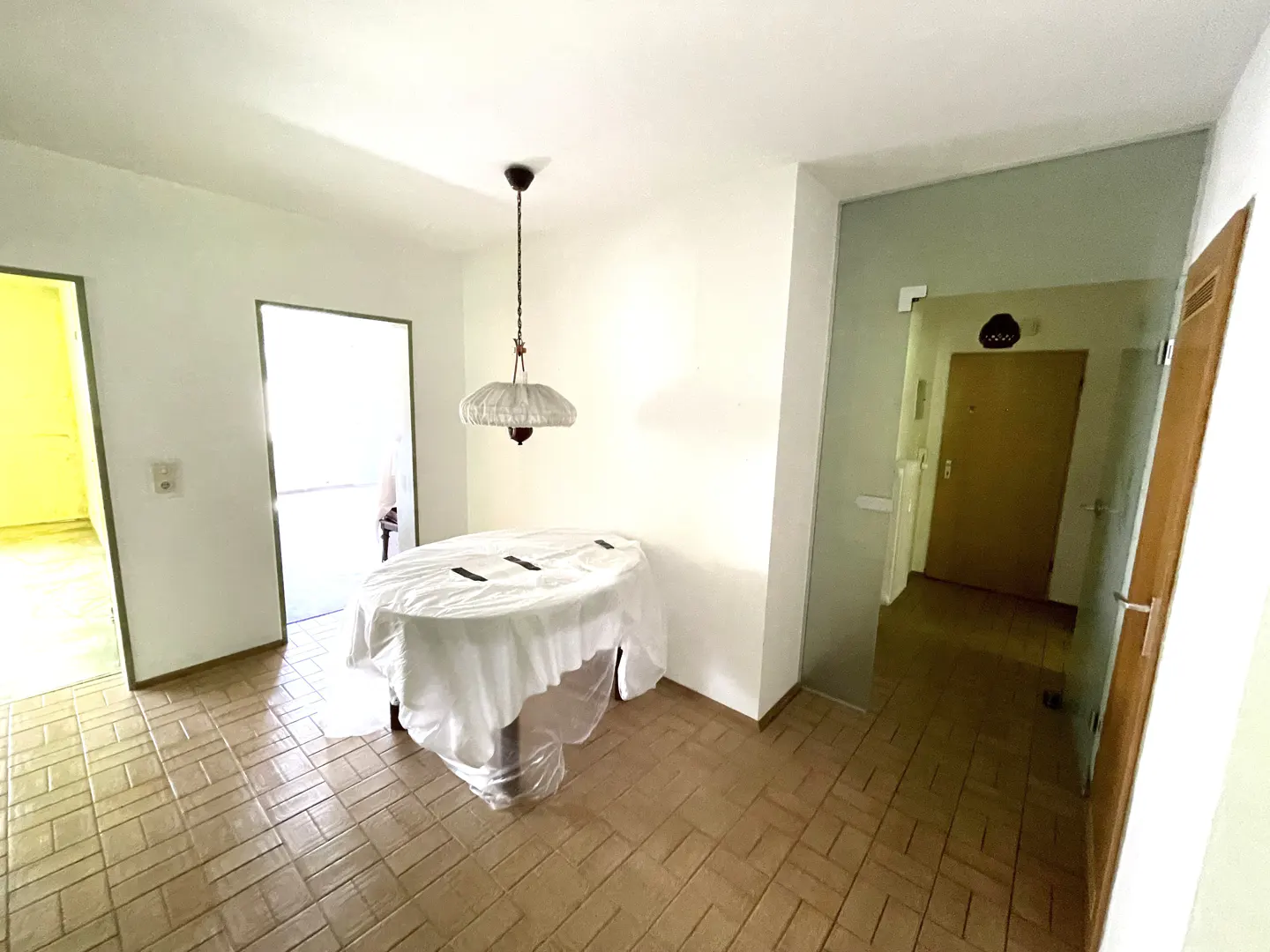 Dining area with a table covered in a white plastic sheet, a hanging pendant light, and tiled floors in a white room.