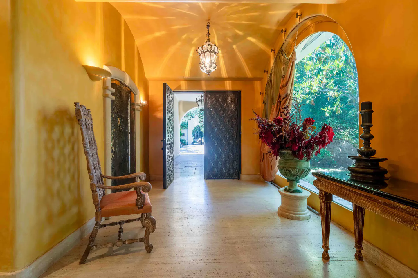 A sunlit Mediterranean-style foyer with yellow walls, a carved wooden chair, a floral urn, and an open arched doorway.