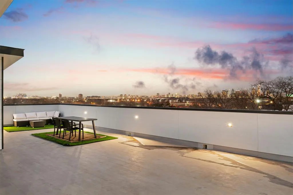 Rooftop patio with white sofa, table, and chairs on artificial turf. City skyline and colorful sunset in the background.