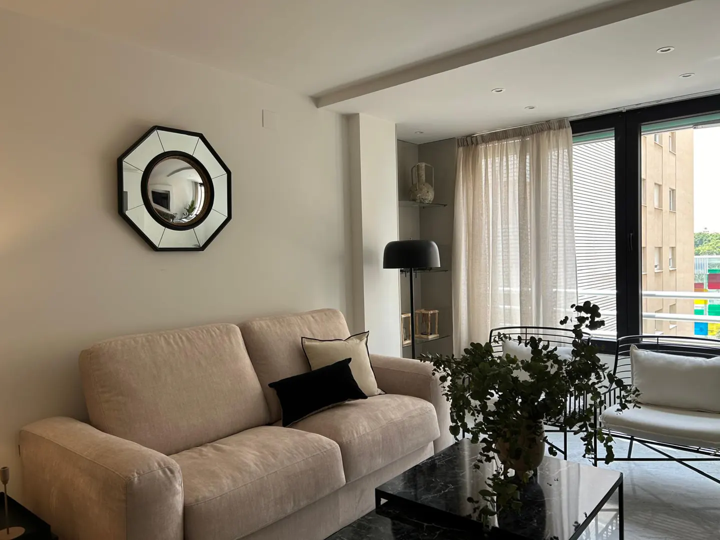 Living room with beige sofa, black pillows, and marble coffee table. Octagon mirror on the wall. Large window with sheer curtains.