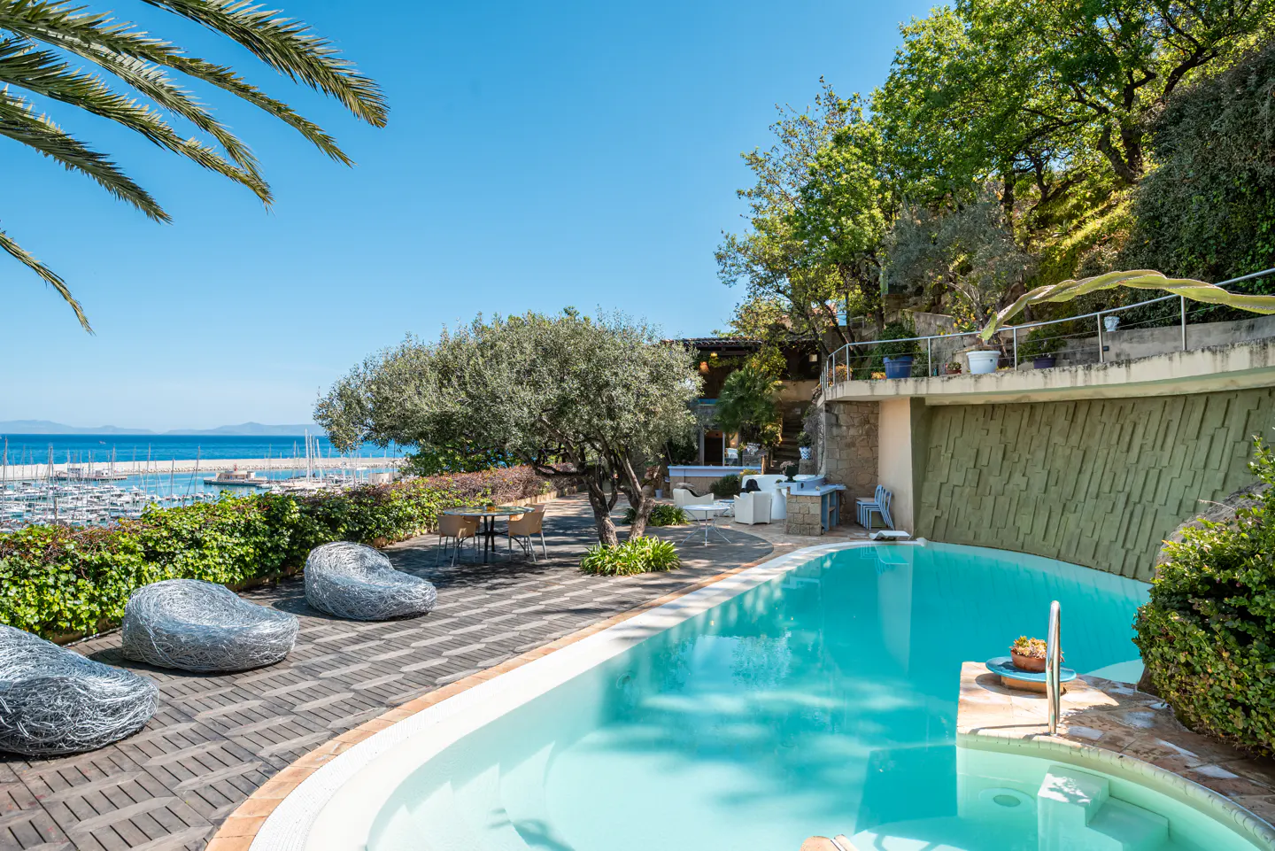 Outdoor pool area with turquoise water, gray stone chairs, and a view of a marina under a clear blue sky.