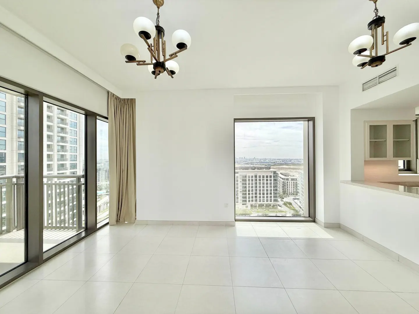 Bright, empty room with white walls, tile floor, and two chandeliers. Large windows offer city views. Beige curtains hang near a balcony.