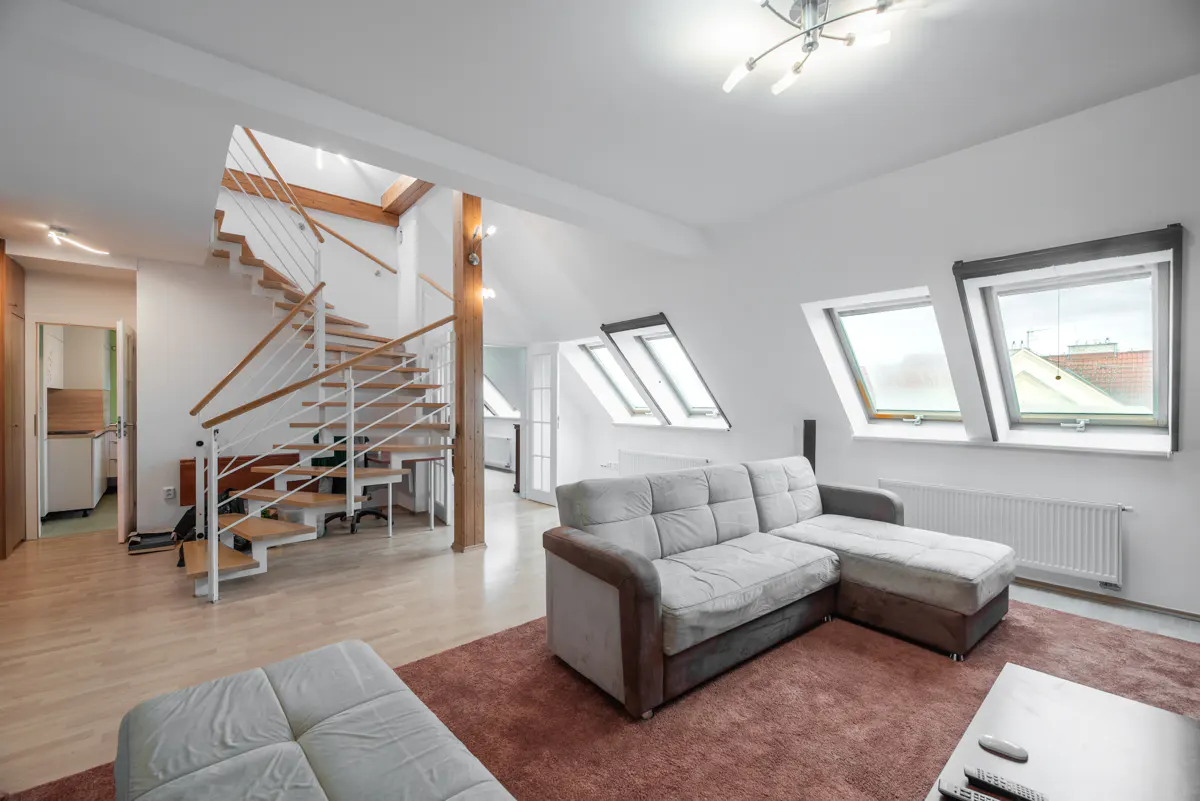 Bright living room with a gray sectional sofa, brown rug, and a modern staircase with white railings. Skylights provide natural light.