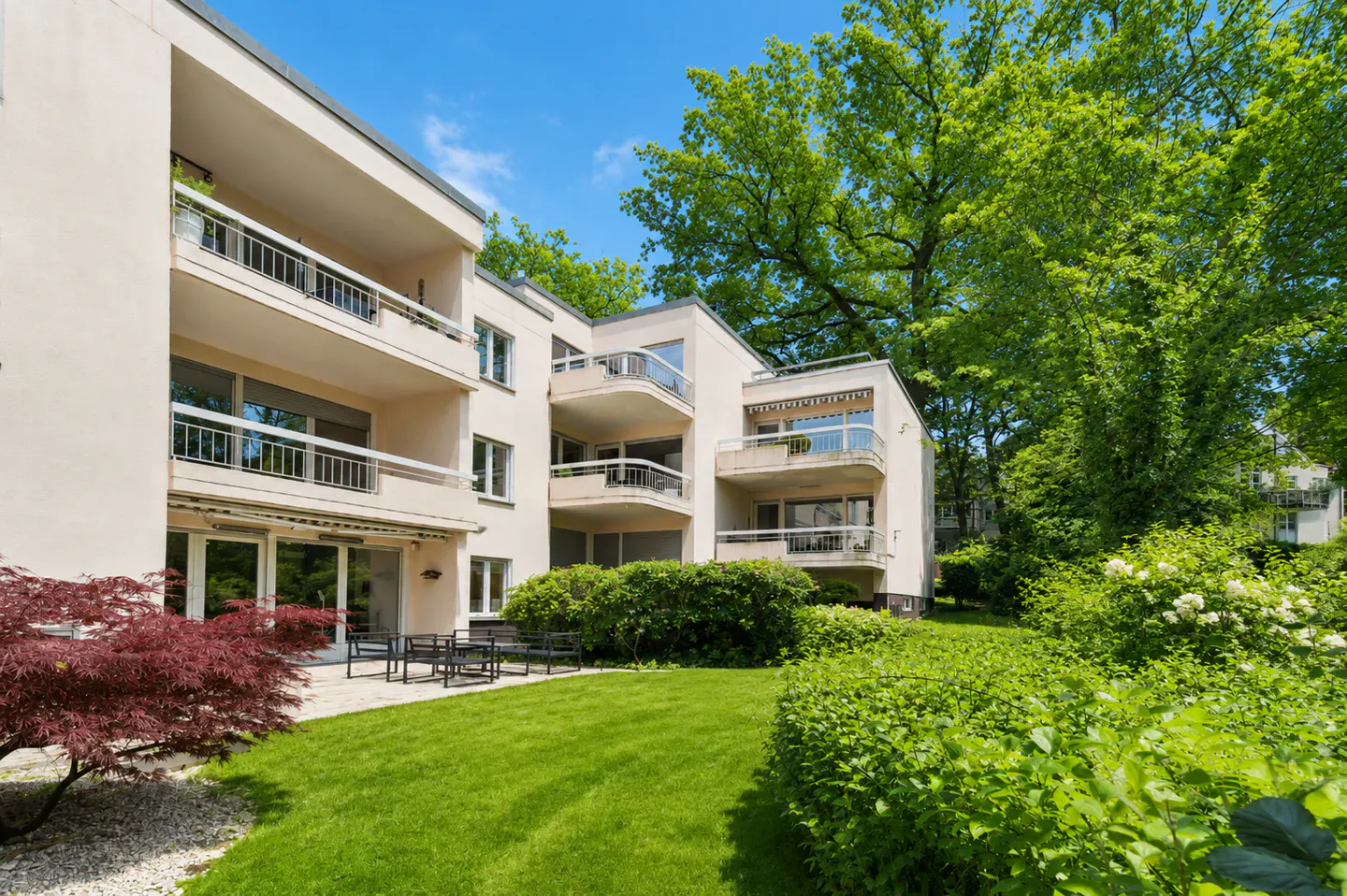 Exterior of a modern, multi-story apartment building with balconies, surrounded by green lawn, trees, and shrubs. Patio furniture sits outside.