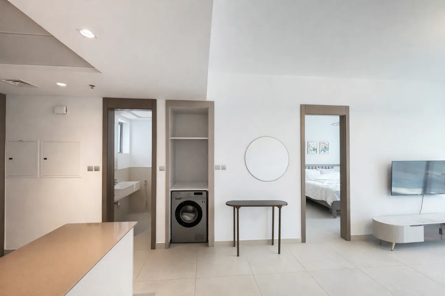 Bright, modern apartment interior with white walls, tile floors, and wood trim. A washing machine is built into a cabinet. A bedroom and bathroom are visible through doorways.