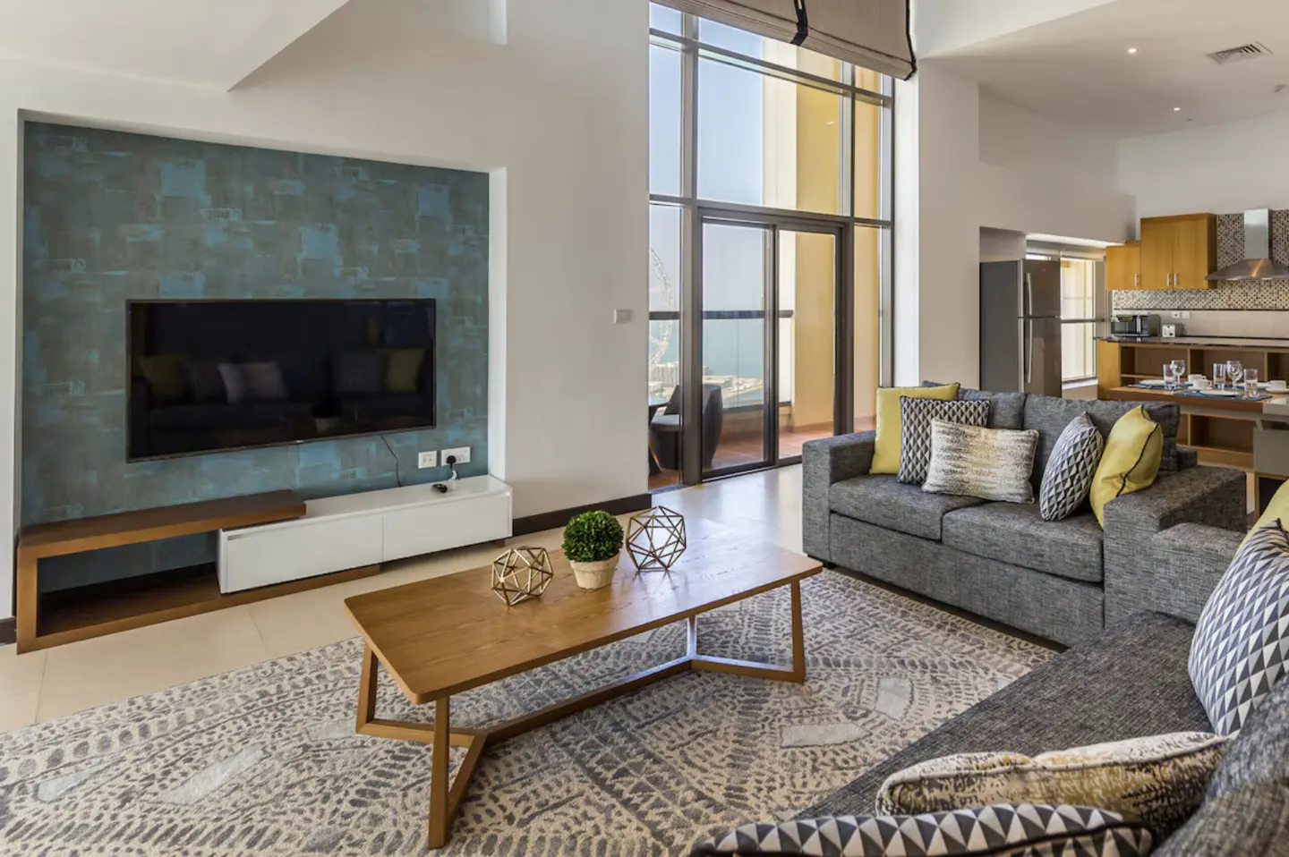 Living room with gray sofas, a wooden coffee table, and a large TV on a blue accent wall. Balcony with ocean view visible through floor-to-ceiling windows.