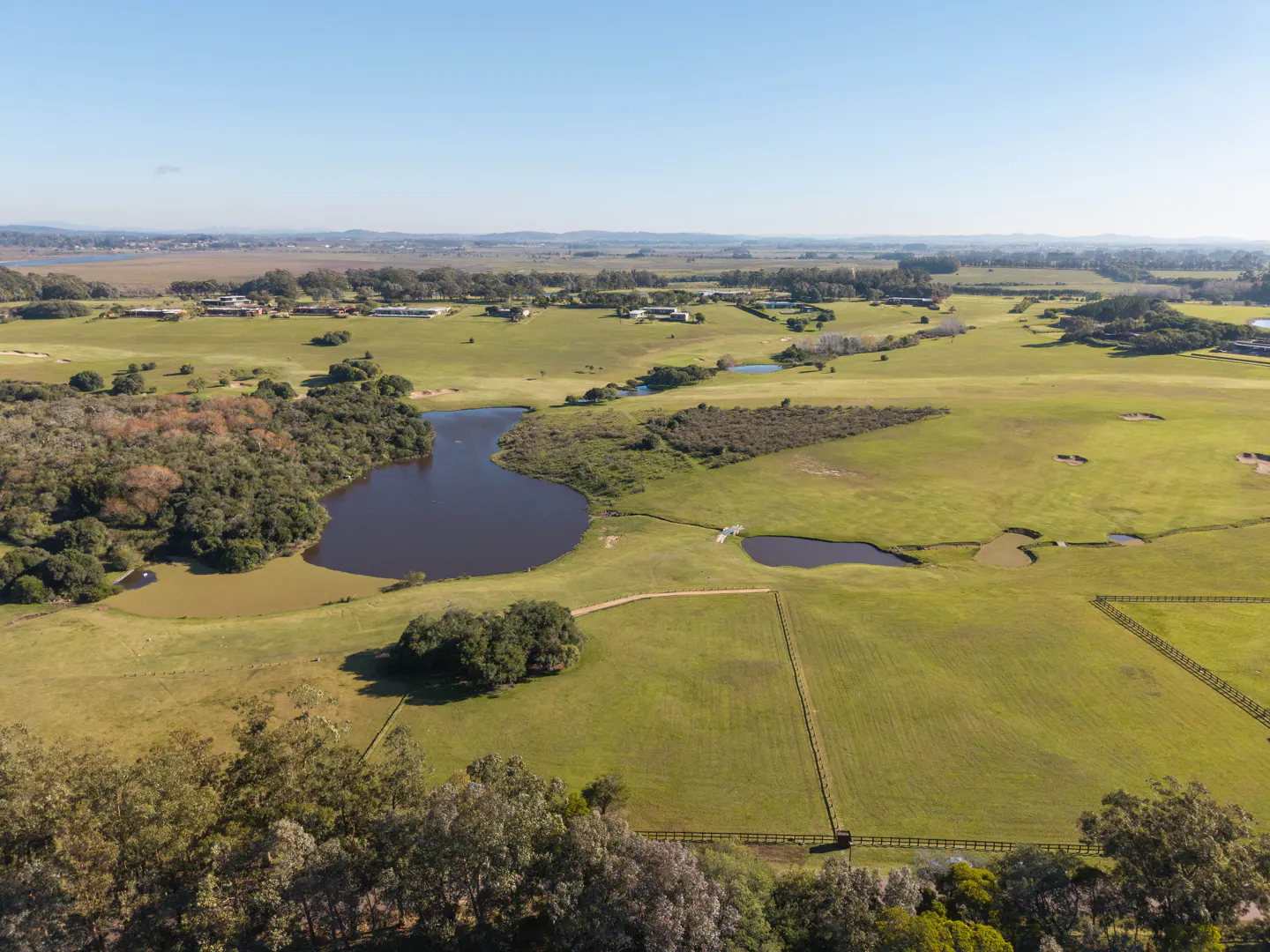 Aerial view of a green pasture with ponds, trees, and fences under a blue sky.