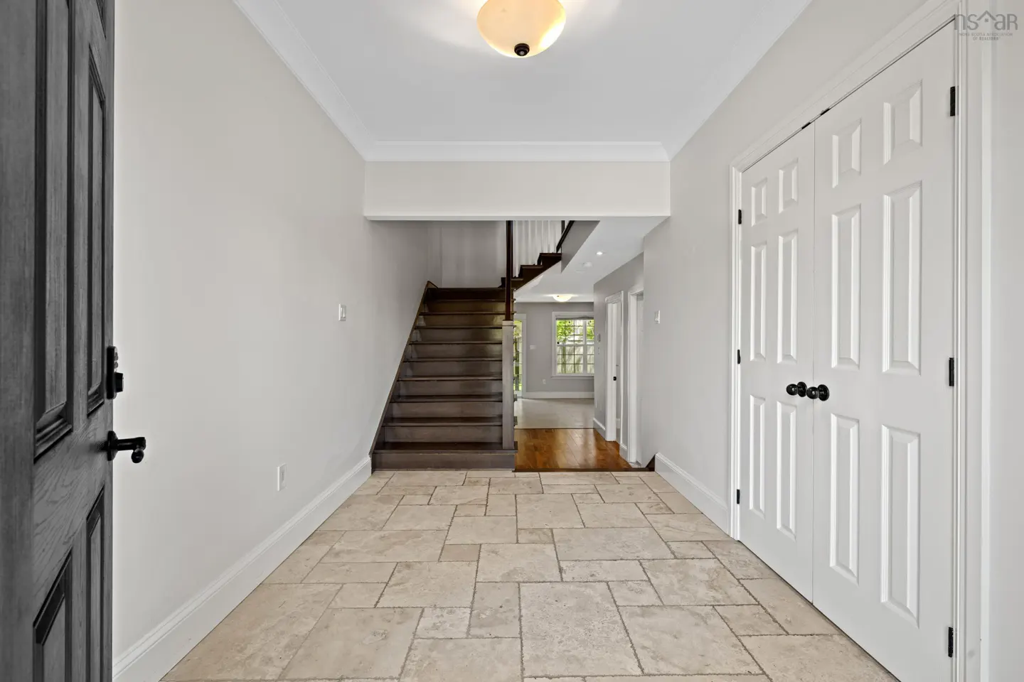 Bright foyer with light stone tile flooring, white walls, a dark wood staircase, and double white closet doors.