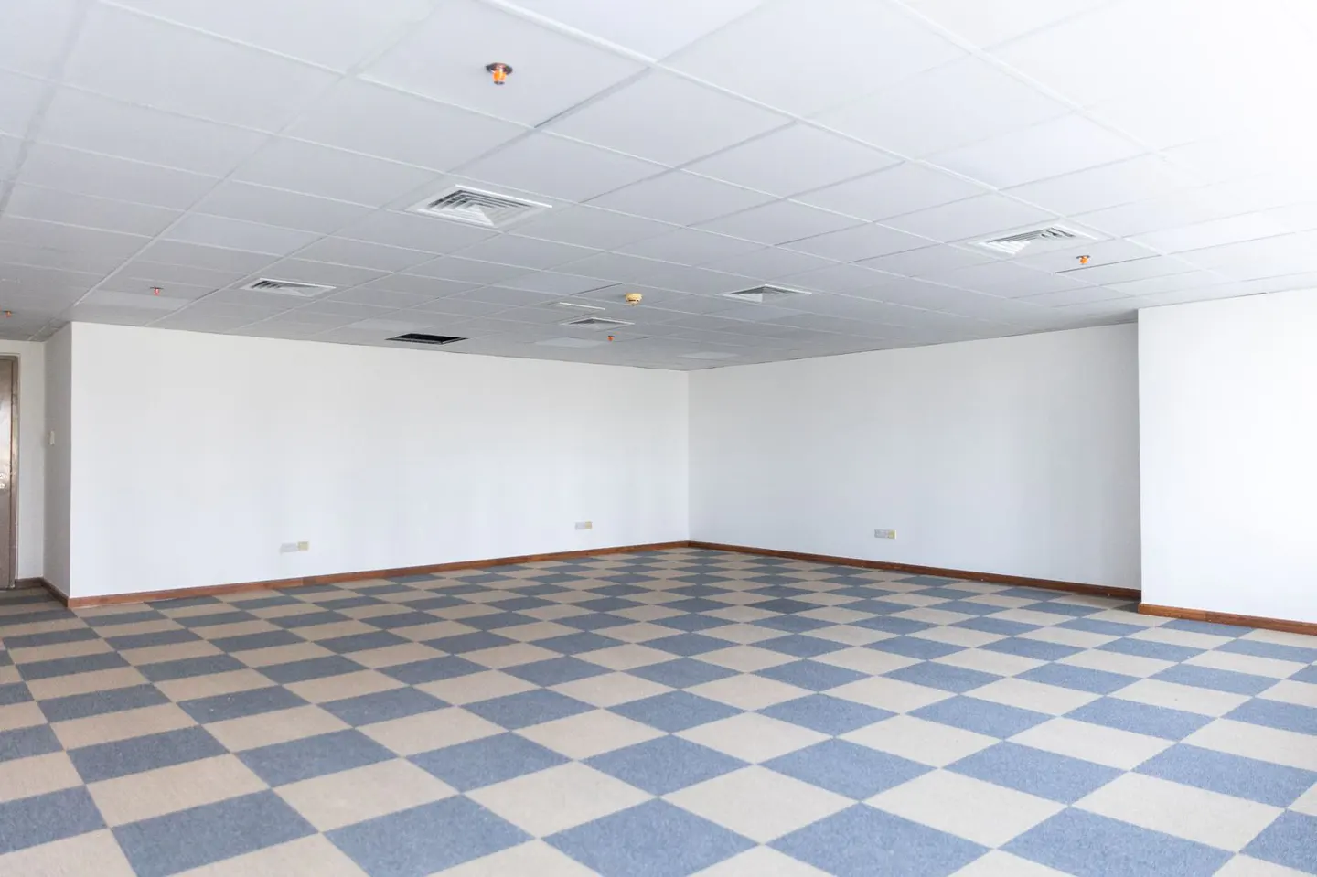 Empty office space with white walls, a checkered blue and beige carpet, and a white tiled ceiling with vents and sprinklers.