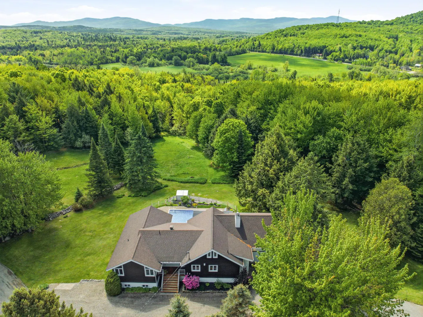 Aerial view of a dark brown house with a pool, surrounded by green trees and mountains in the background.