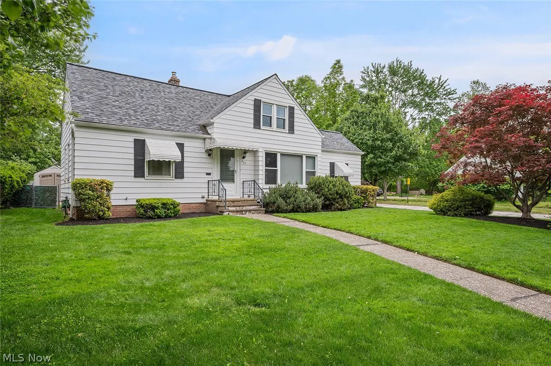 Exterior view of a white, one-story house with black shutters and a gray roof, surrounded by a green lawn and trees.