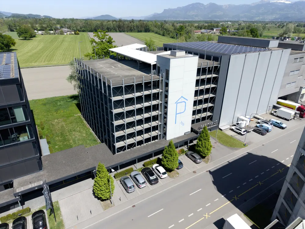 Aerial view of a modern parking garage with solar panels, cars parked outside, and green fields in the background.