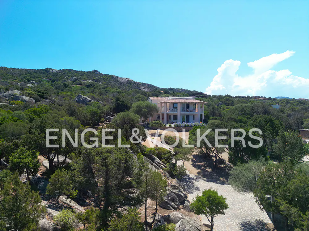 A multi-story villa with a tiled roof nestled in a lush, rocky hillside landscape under a clear blue sky.
