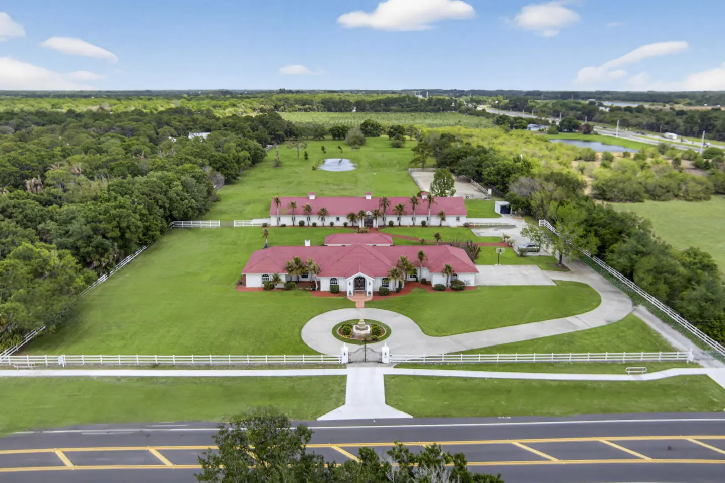 Aerial view of a white house with a red roof, surrounded by green grass and trees. A circular driveway leads to the entrance.