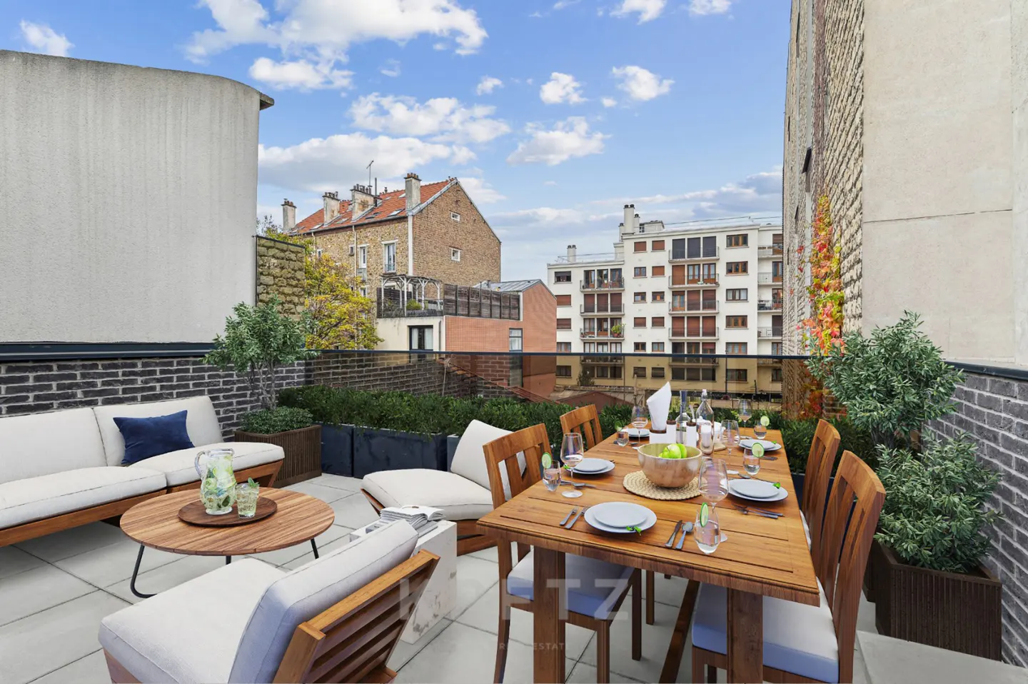 Rooftop terrace with a wooden dining set, white lounge seating, potted plants, and city buildings under a blue sky.