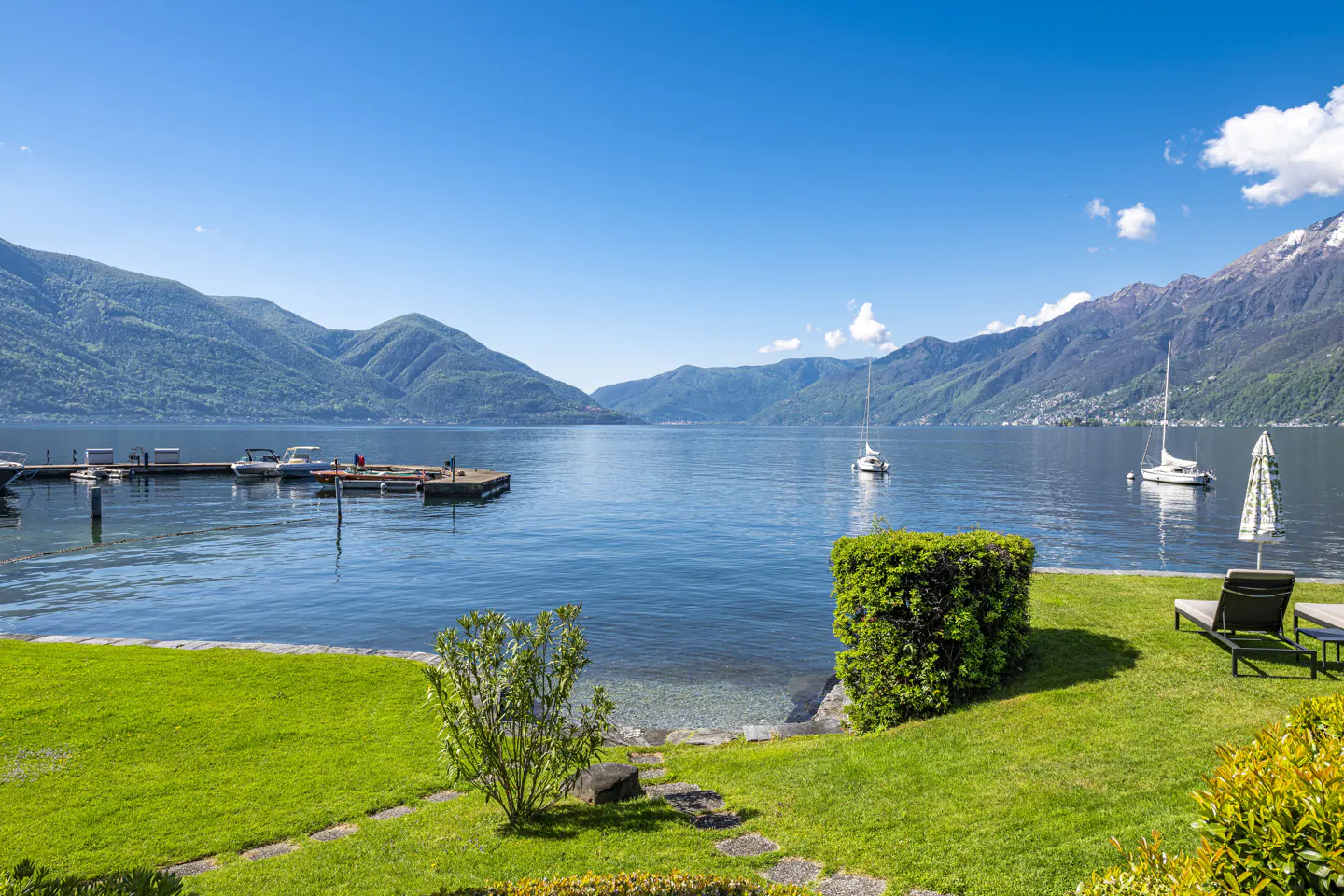 Scenic lake view with mountains, boats, and green lawn. Blue sky above. Deck chairs on the grass.