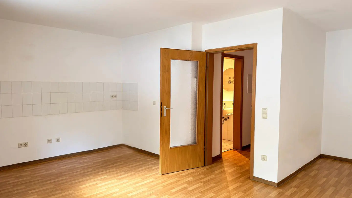 A bright, empty room with wood floors, white walls, and an open wood door leading to a bathroom.