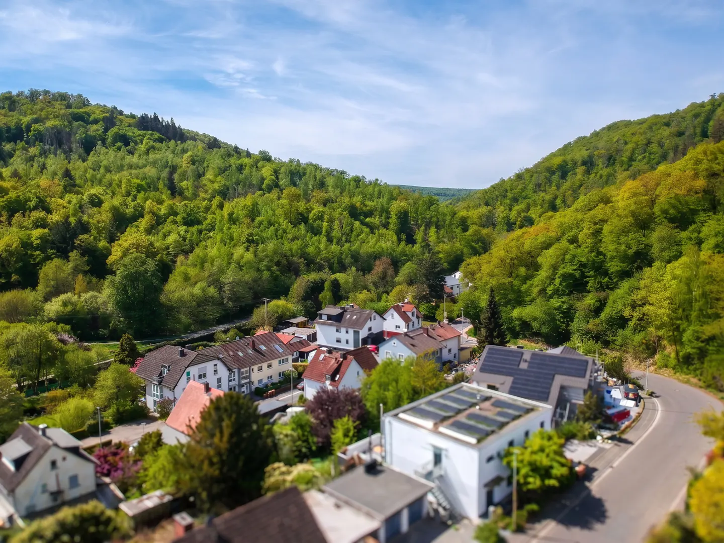 Aerial view of a village nestled in a green valley, with houses, trees, and a winding road under a blue sky.