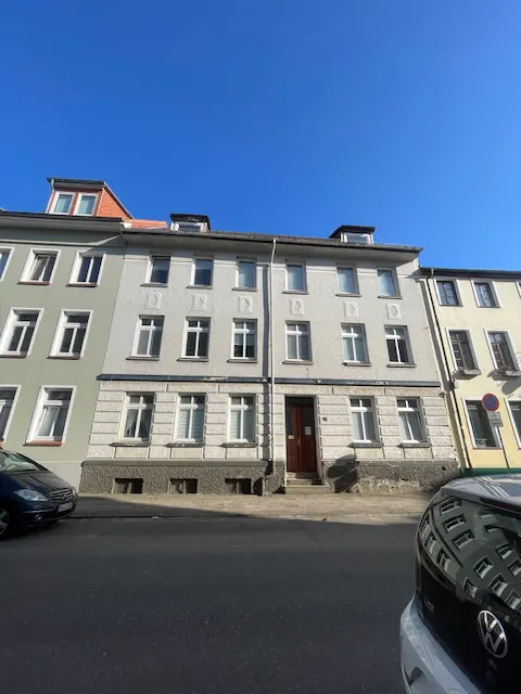 Three-story apartment building with white windows and a brown door under a clear blue sky. Cars are parked on the street.