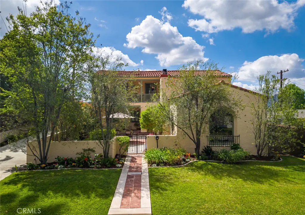 Two-story beige house with a red tile roof, green lawn, trees, and a brick walkway under a blue sky with white clouds.