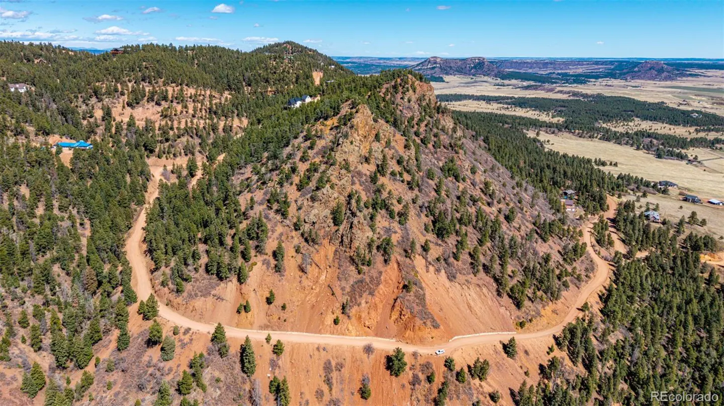 Aerial view of a mountain with a dirt road winding around it, surrounded by green trees and a blue sky.