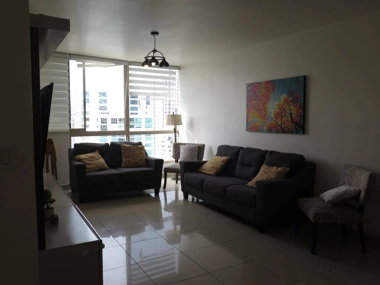 Living room with two gray sofas, patterned chairs, and a colorful painting. Large window with blinds shows a city view.
