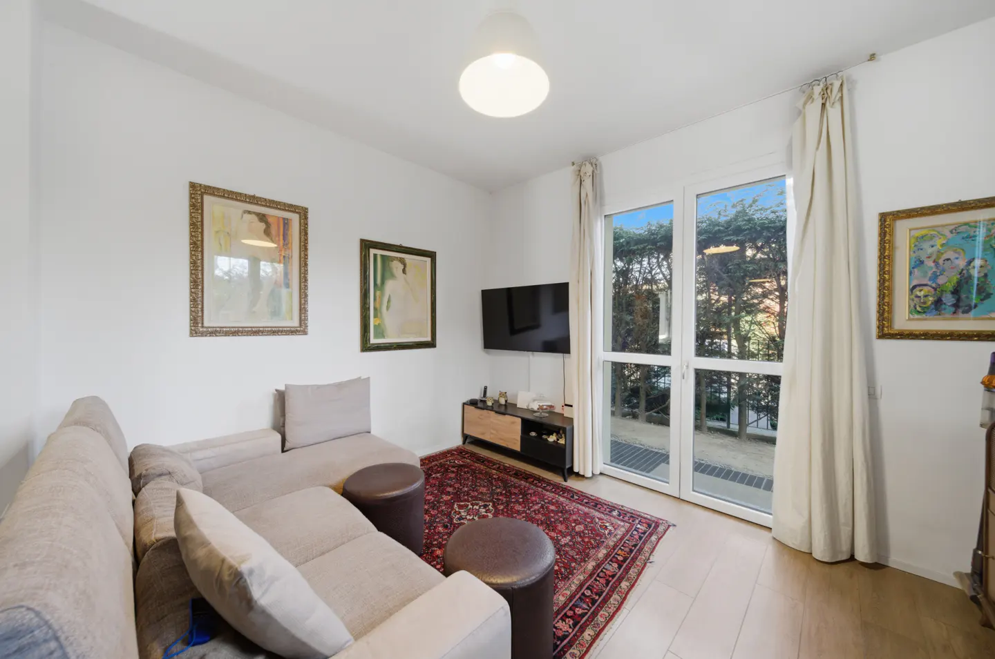Bright living room with a beige sectional sofa, two brown ottomans, and a red patterned rug. A TV hangs above a wooden console table. Large windows offer a view of trees.