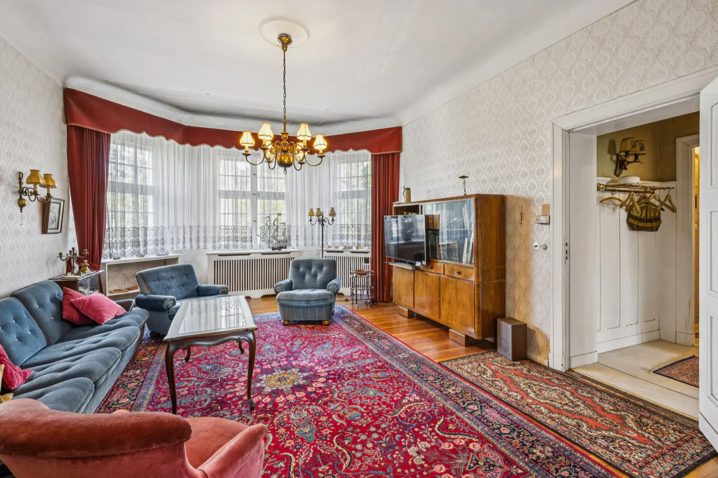 Living room with blue sofas, red rug, wood floors, and a large window with red curtains. A chandelier hangs from the ceiling.