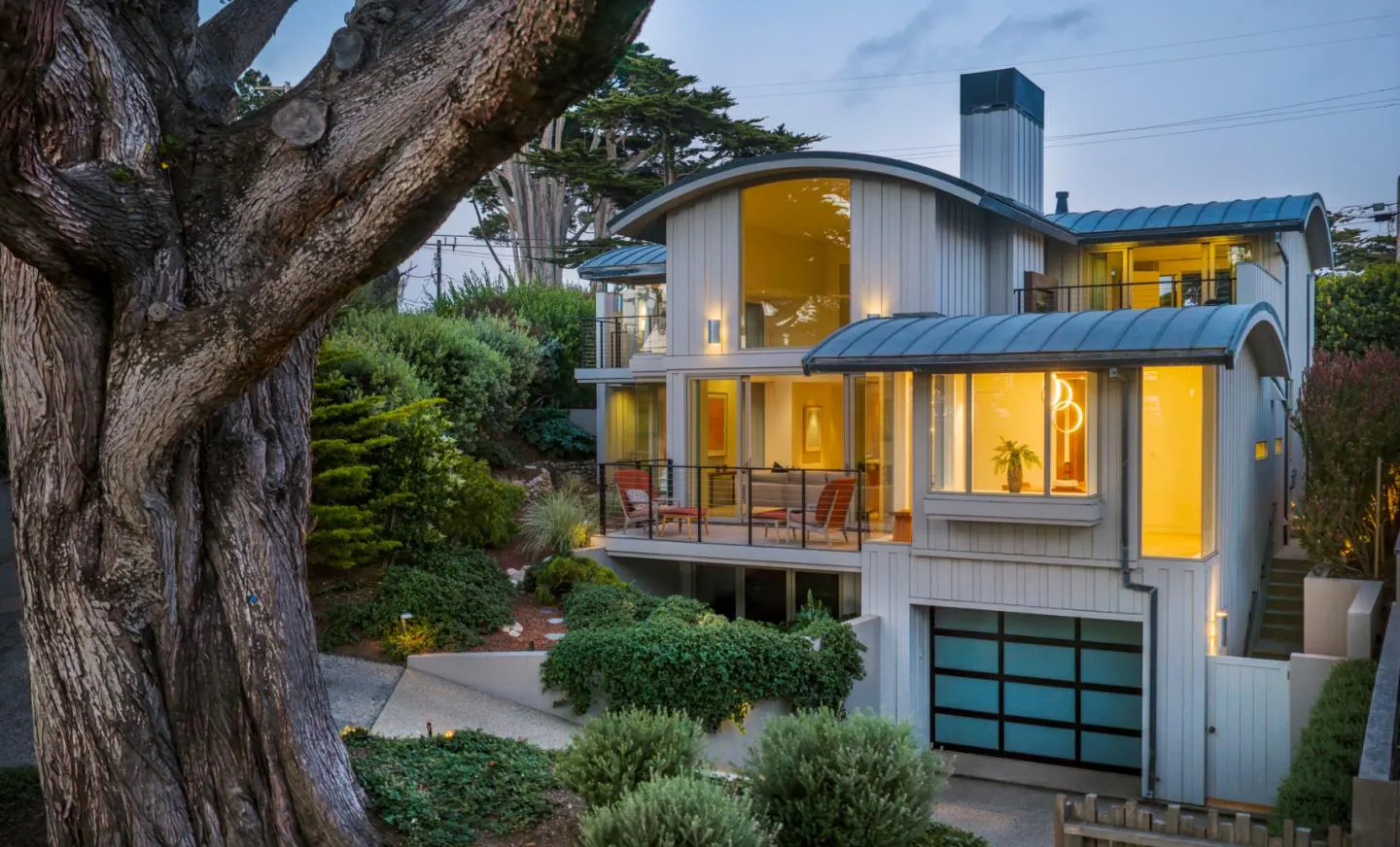 Modern multi-level home with curved roof, illuminated windows, and a garage, set in a lush, tree-filled landscape at dusk.