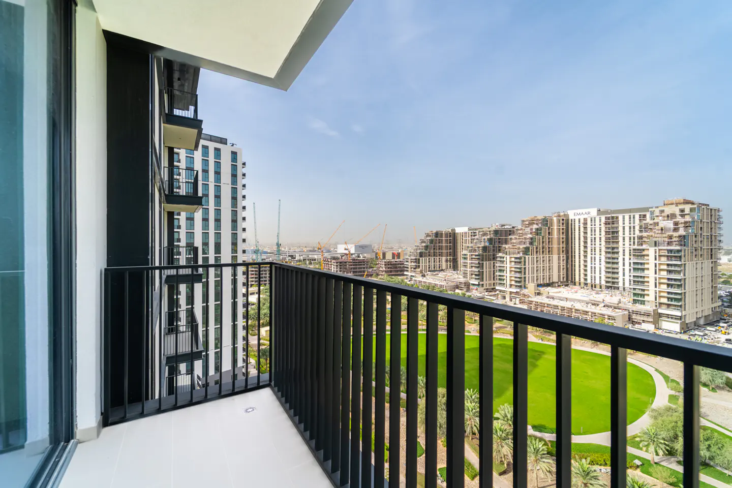 View from a balcony with black railings overlooking a green park and city buildings under a blue sky.