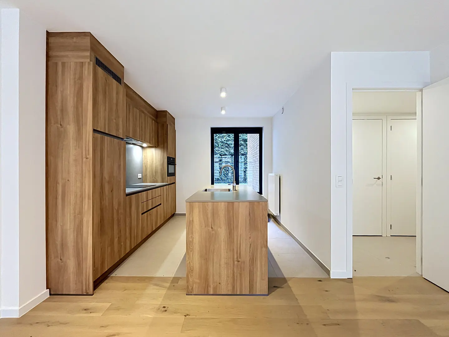 A bright, modern kitchen with wood cabinets, a center island, and stainless steel appliances. White walls and light wood floors create a clean, airy feel.