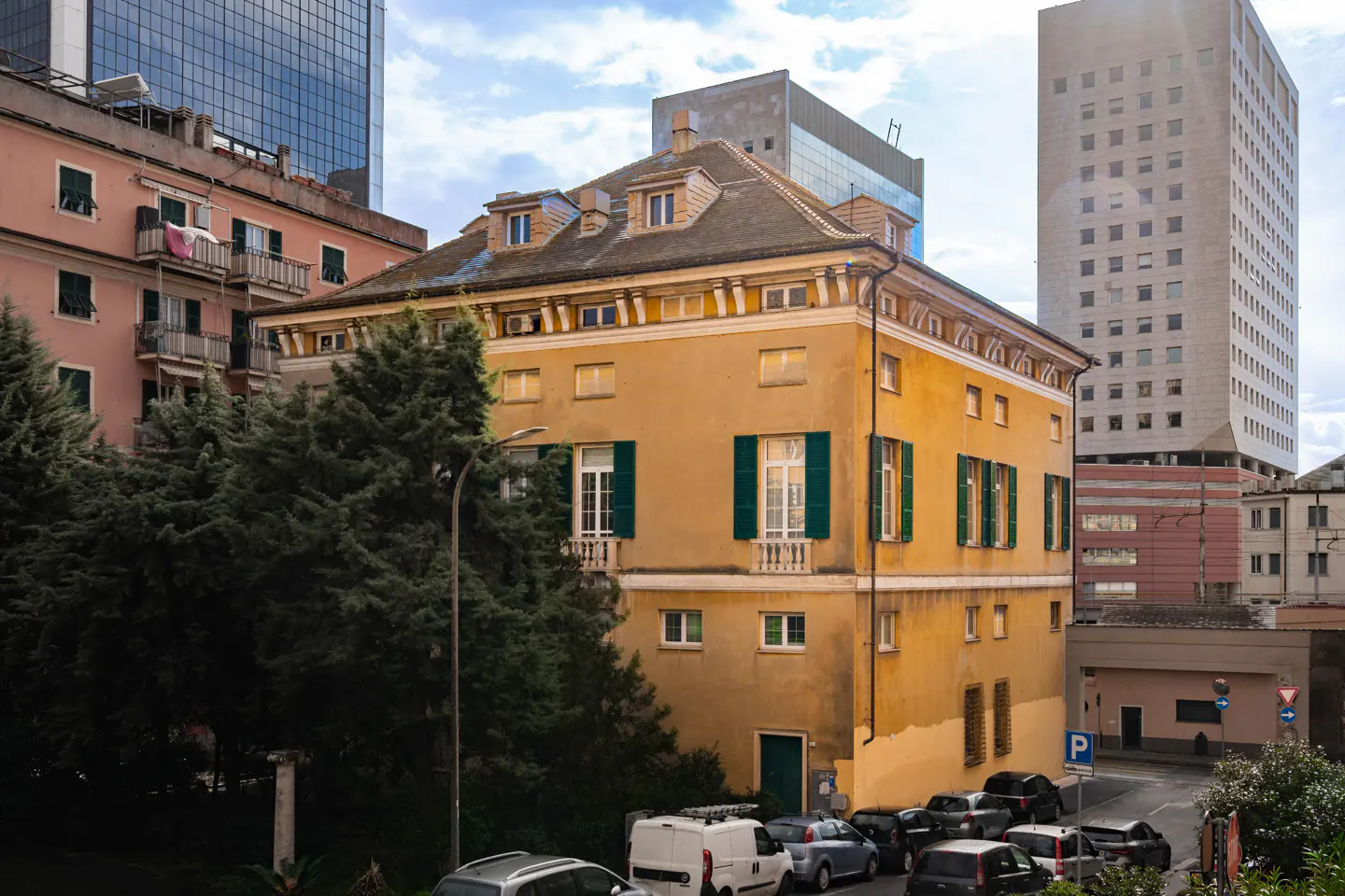A three-story yellow building with green shutters, surrounded by trees and modern buildings.