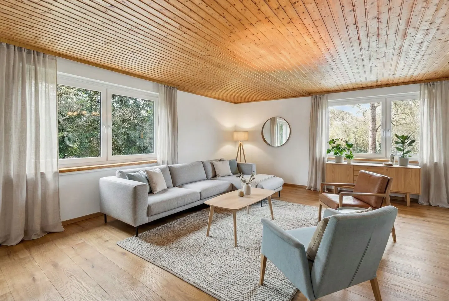 Living room with wood ceiling, light gray sectional sofa, two chairs, and large windows with sheer curtains.