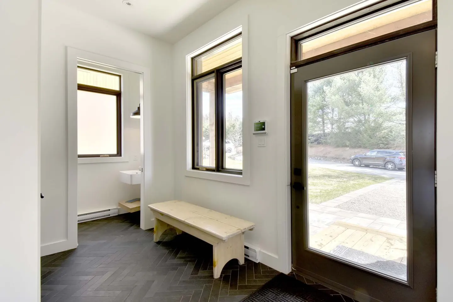 Bright entryway with black herringbone floors, white walls, and a wooden bench under a window. A dark door leads outside.