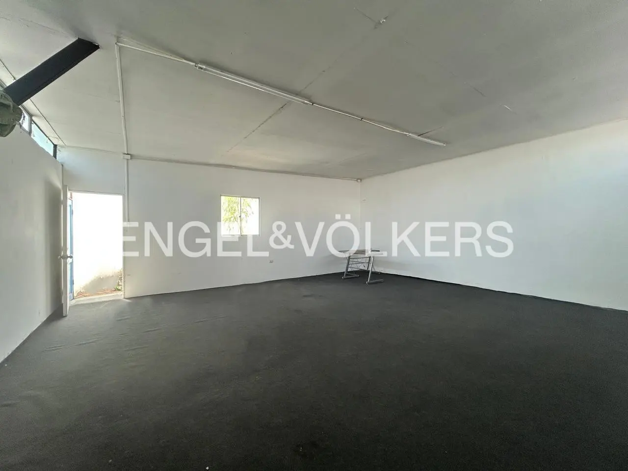 Empty commercial space with black floor, white walls, and a small window. An open door is on the left. Engel & Völkers logo is visible.