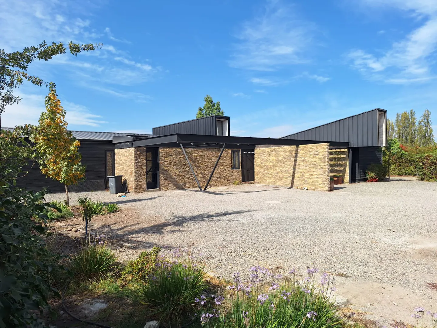 Modern home with stone and dark wood exterior, black metal roof, gravel driveway, and blue sky.