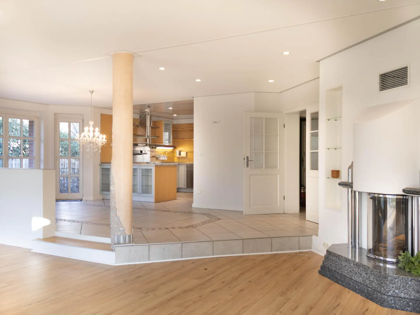 Open-concept living space with wood floors, tile transition, and a view into the kitchen. A chandelier hangs near large windows.