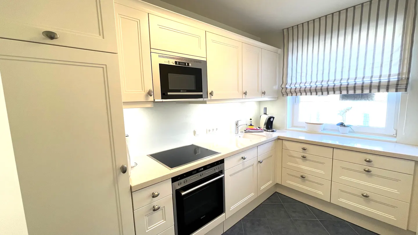 A bright kitchen with white cabinets, a black stovetop and oven, and a microwave. A window with striped curtains is above the counter.