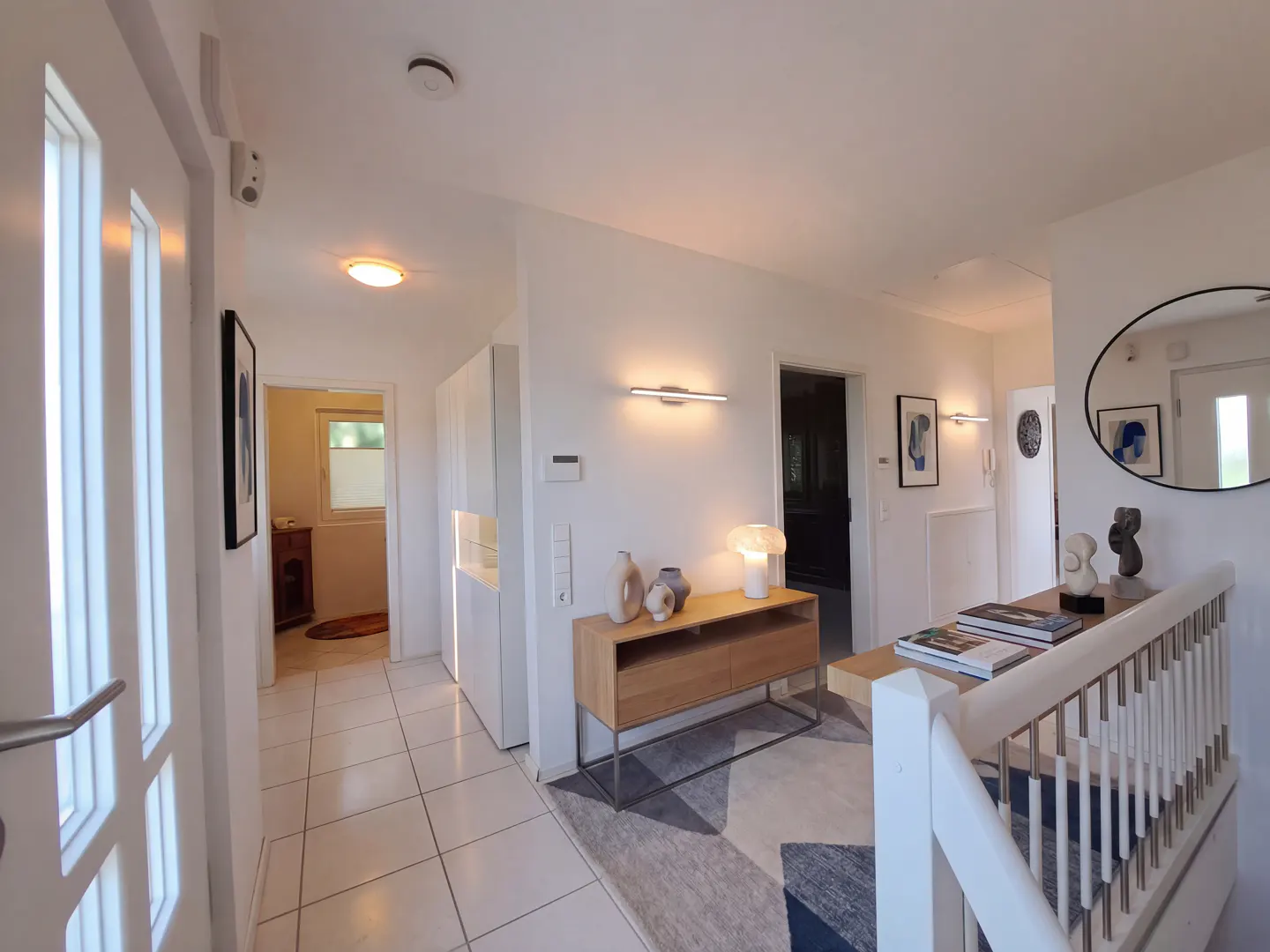 Bright foyer with white walls, tiled floor, and natural wood accents. A staircase with white railings is visible on the right.